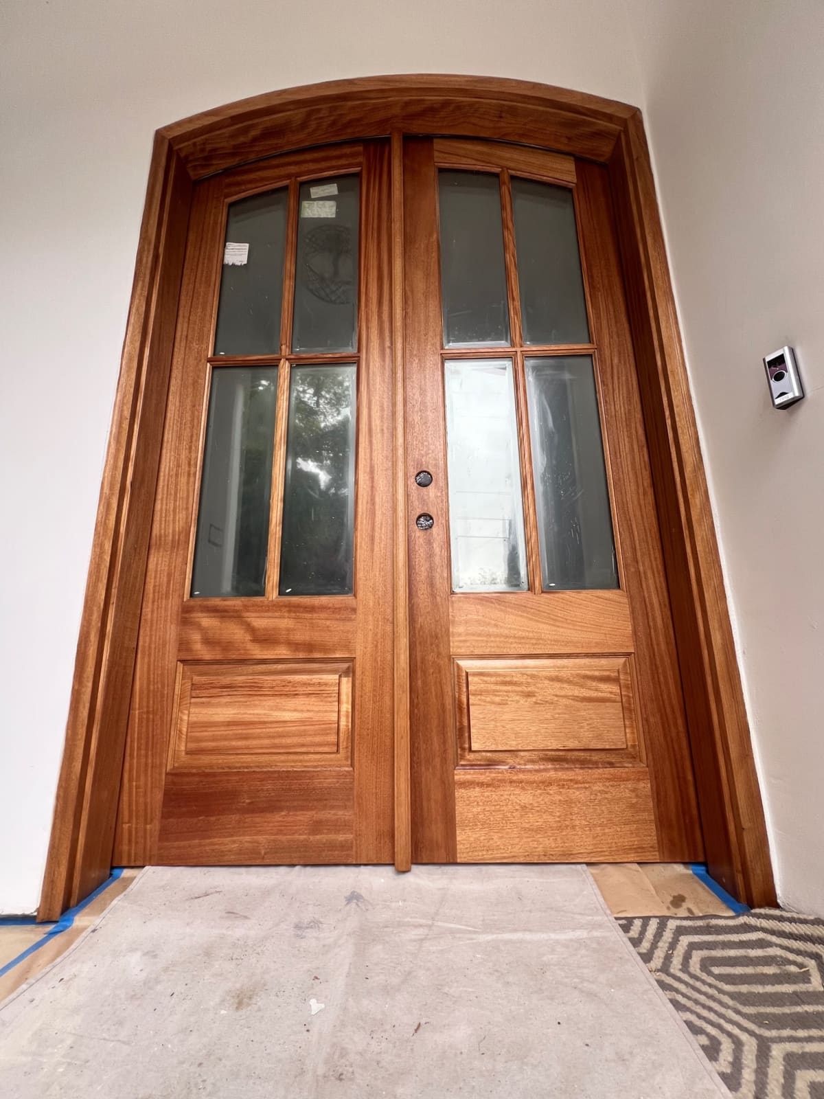 Arched mahogany double front door before and after staining — from raw, unfinished wood to a rich, deep wood-grain finish